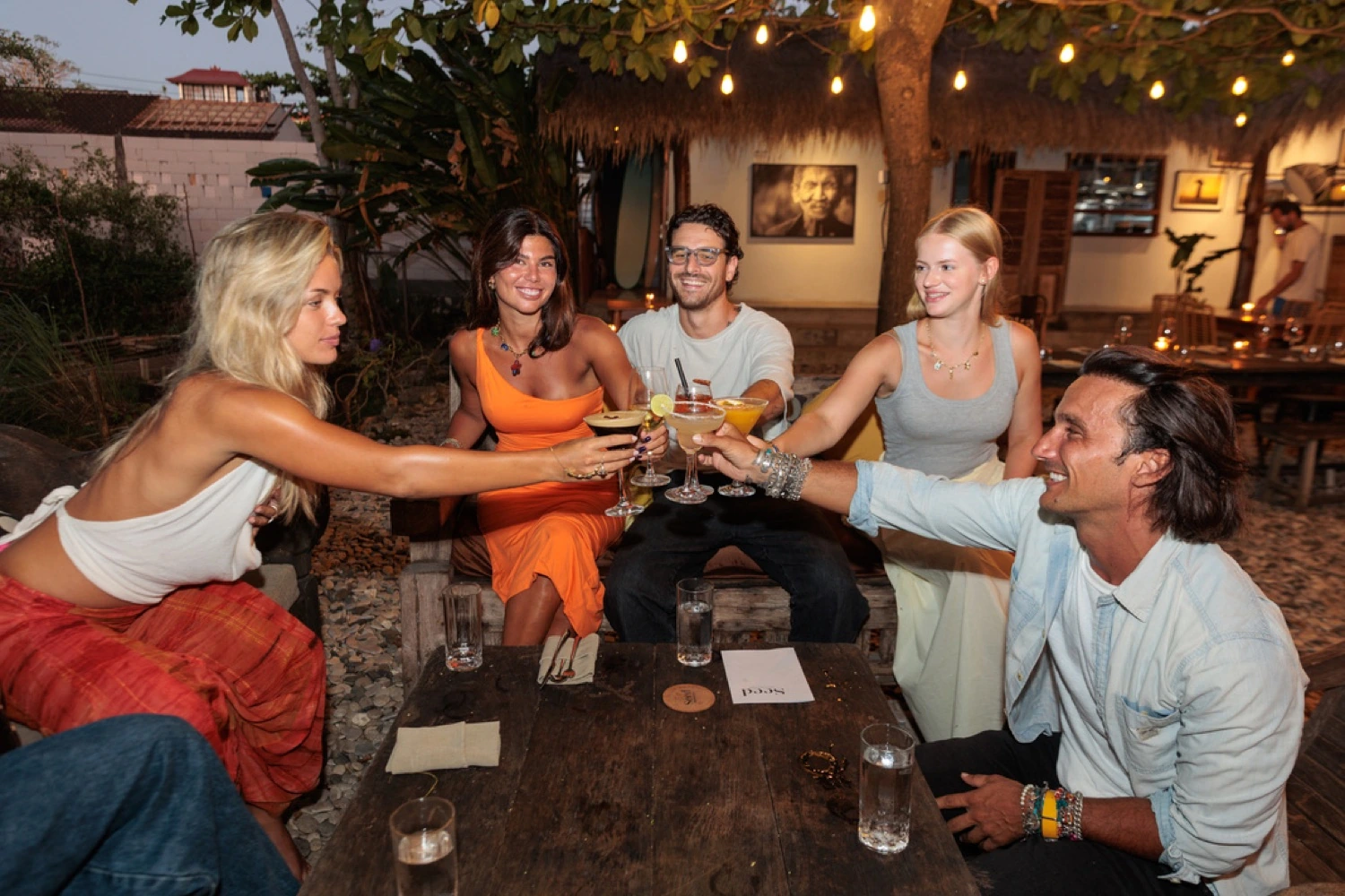 Five guests raising cocktails around a garden table at Seed in Bingin, under string lights at dusk