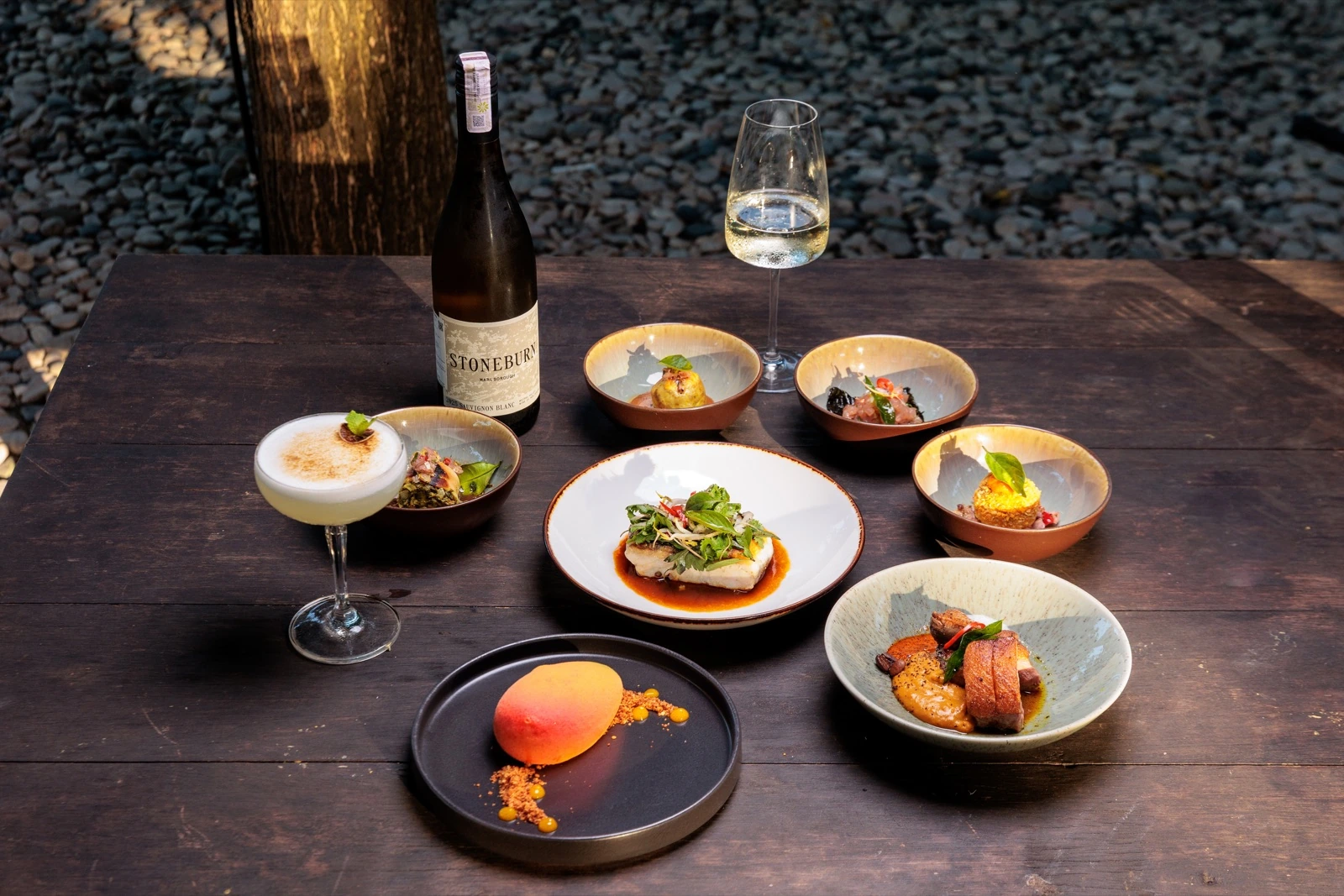 The seven course Nusantara tasting menu laid out on a wooden table, small plates, a ginger parfait, a cocktail and a bottle of white wine.