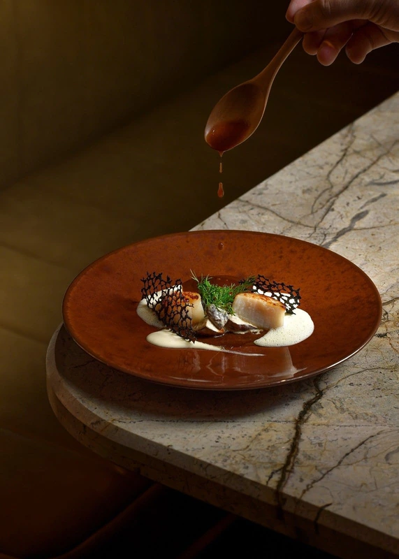 Scallops with a dill garnish and a spoon of sauce poured over, plated on a copper dish at Seed