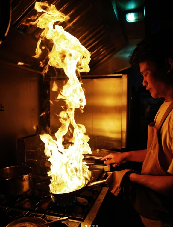 A cook flaming a pan, the fire rising high above the stove in the Seed kitchen