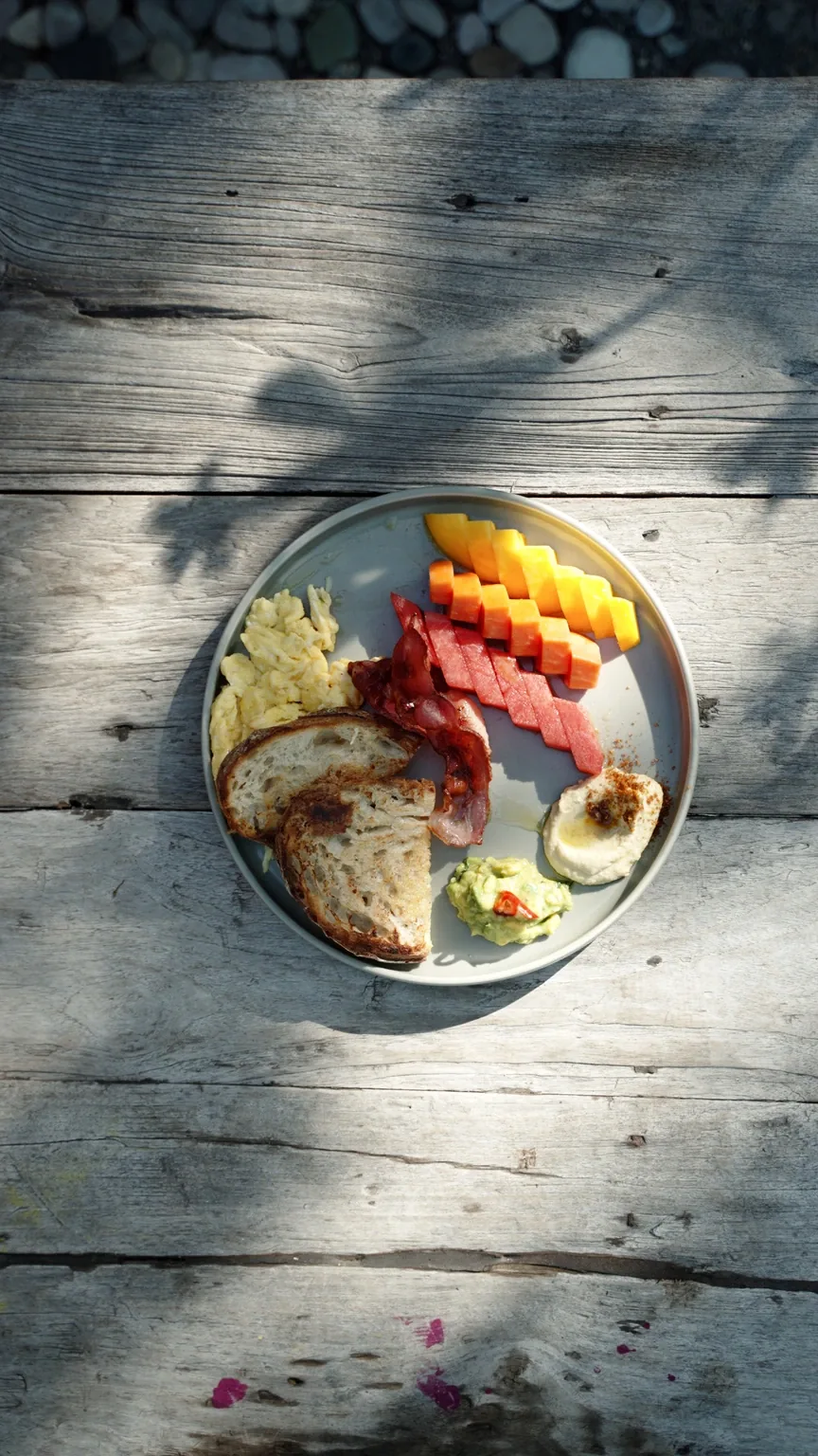 A breakfast plate of scrambled egg, bacon, sourdough, hummus and fruit on a sunlit table at Seed